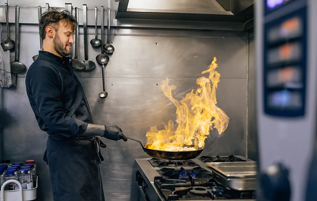 Un chef en tenue noire fait flamber des ingrédients dans une poêle sur une cuisinière professionnelle en acier inoxydable.